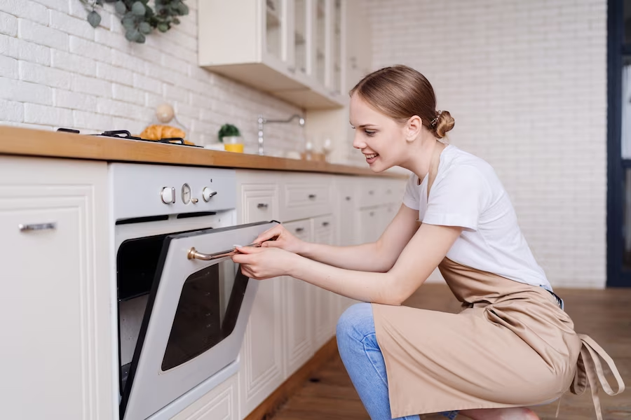 A girl in the kitchen wearing an apron, opening an oven